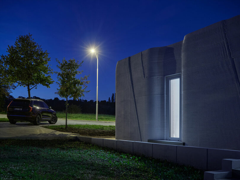 A car is parked near trees and a streetlight on a road at night, with a modern, textured building in the foreground.