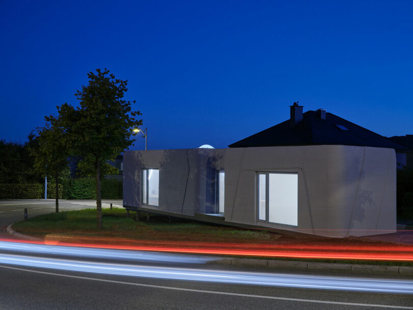 A modern, minimalist house with illuminated windows sits near a road at dusk; light trails from passing cars are visible in the foreground.