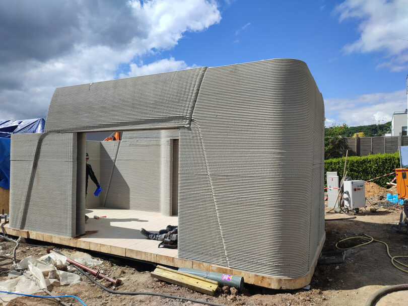 A partially constructed house with curved walls made from layered concrete, built using 3D printing technology, stands on a construction site under a partly cloudy sky.