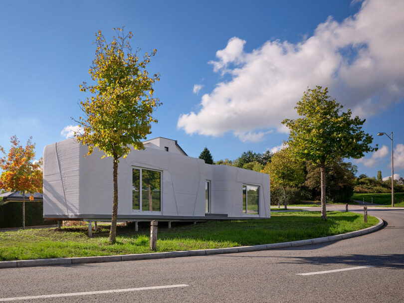 Modern white house with large windows sits on a grassy corner lot near the street, surrounded by a few trees under a blue sky with scattered clouds.