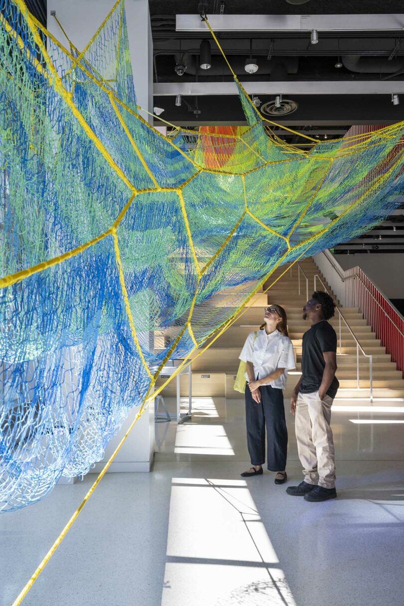 Two people stand beneath a large, colorful net-like installation suspended from the ceiling in a modern, sunlit gallery space.