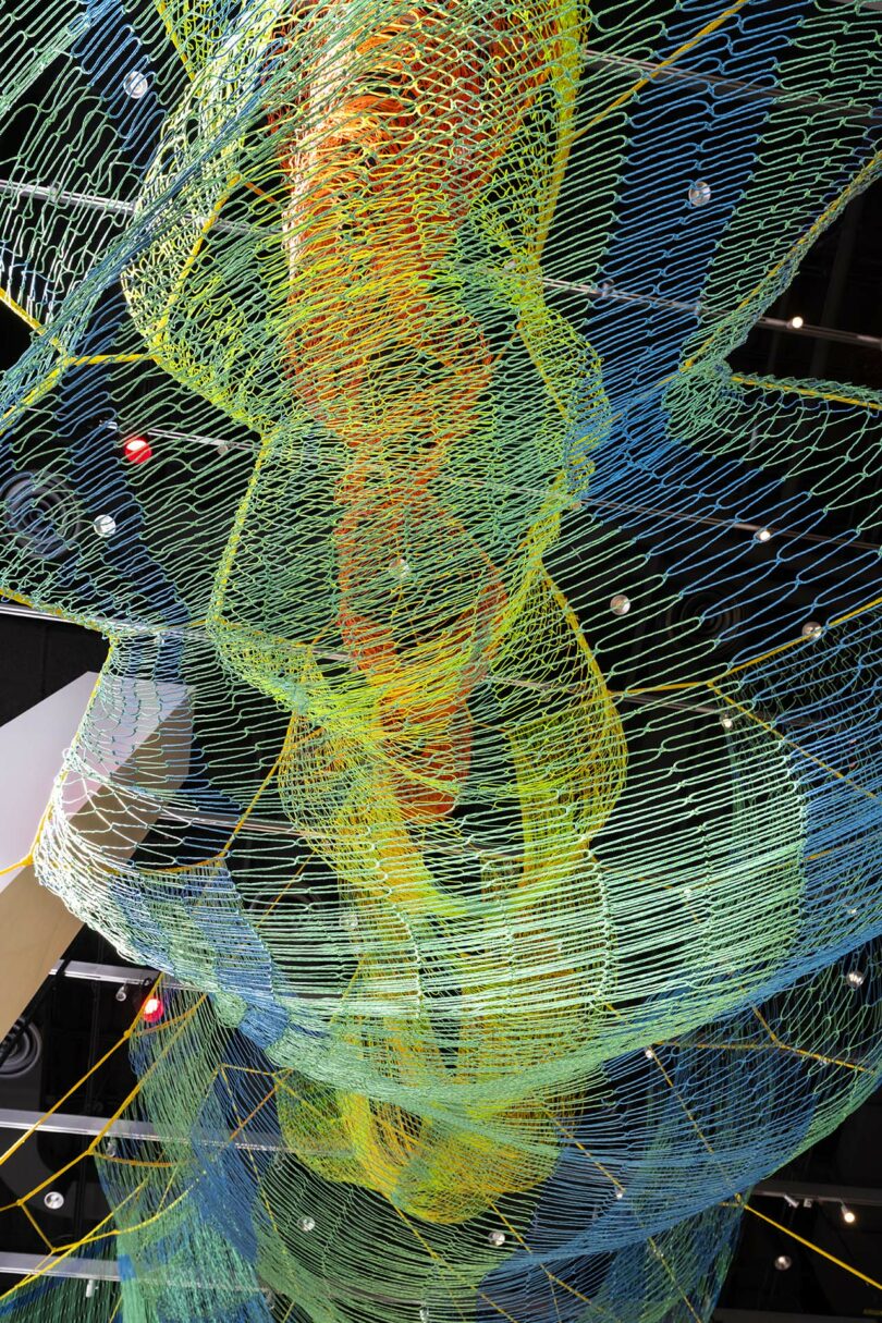 Colorful woven net sculpture with blue, green, and yellow hues hangs from the ceiling, viewed from below, with lights and dark ceiling panels in the background.