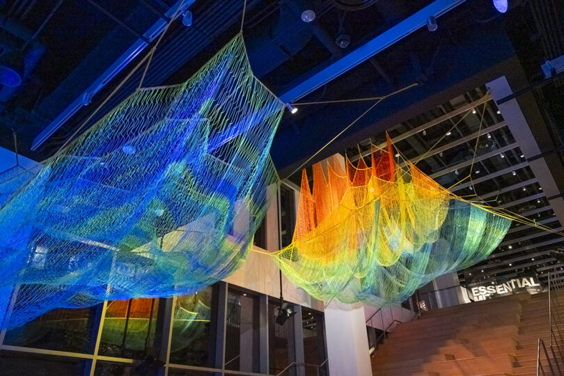 Colorful net-like installation art suspended from the ceiling, transitioning from blue to green to orange, above a staircase in a modern interior space.