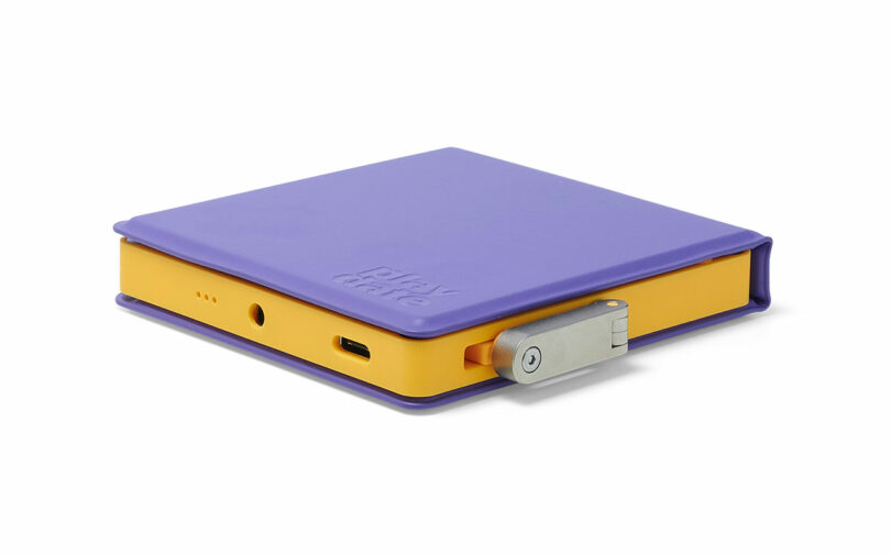 A compact, square electronic device with a purple cover, yellow sides, a metal latch, and ports for audio and USB connections. The word