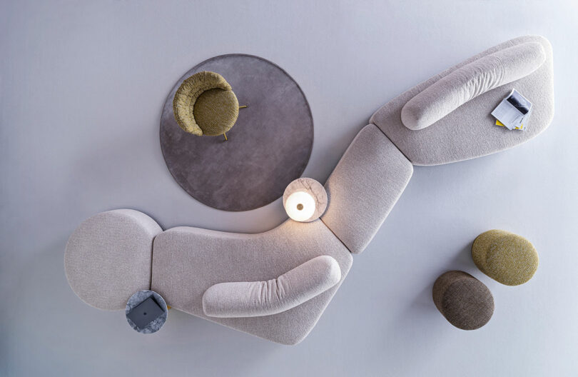 A top-down view of modern, modular beige sofas, a round gray rug, a green chair, two ottomans, a lamp, and a side table with a magazine and notebook.