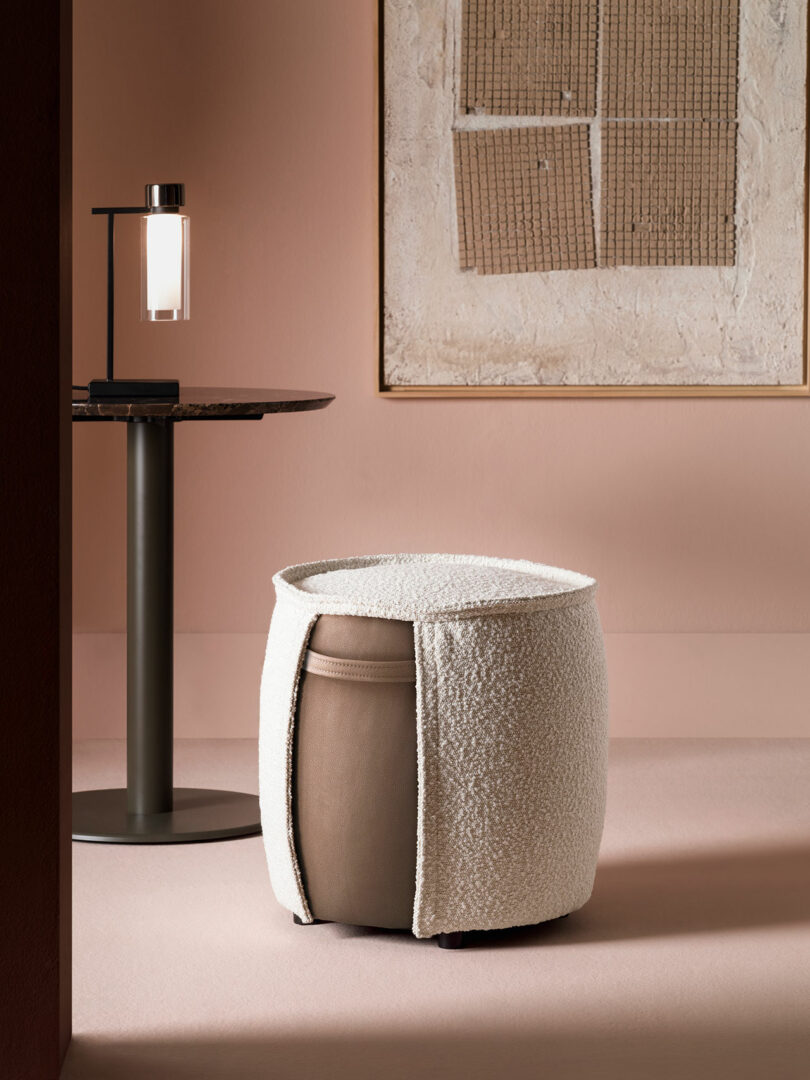 A modern interior with a round side table and lamp, abstract wall art, and a textured, cylindrical ottoman partially covered in white fabric.