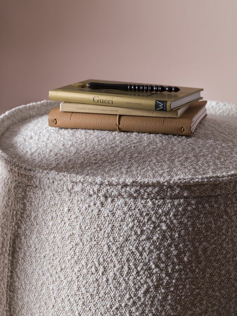 A stack of two books and a closed tan leather notebook with a pen rest on a textured beige upholstered surface against a light pink wall.