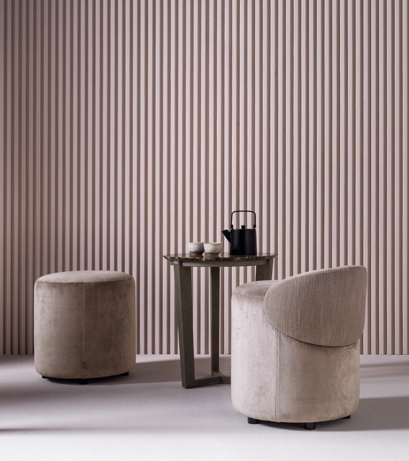 A round ottoman, an armchair, and a small table with a teapot and cups are set against a vertical striped beige wall.