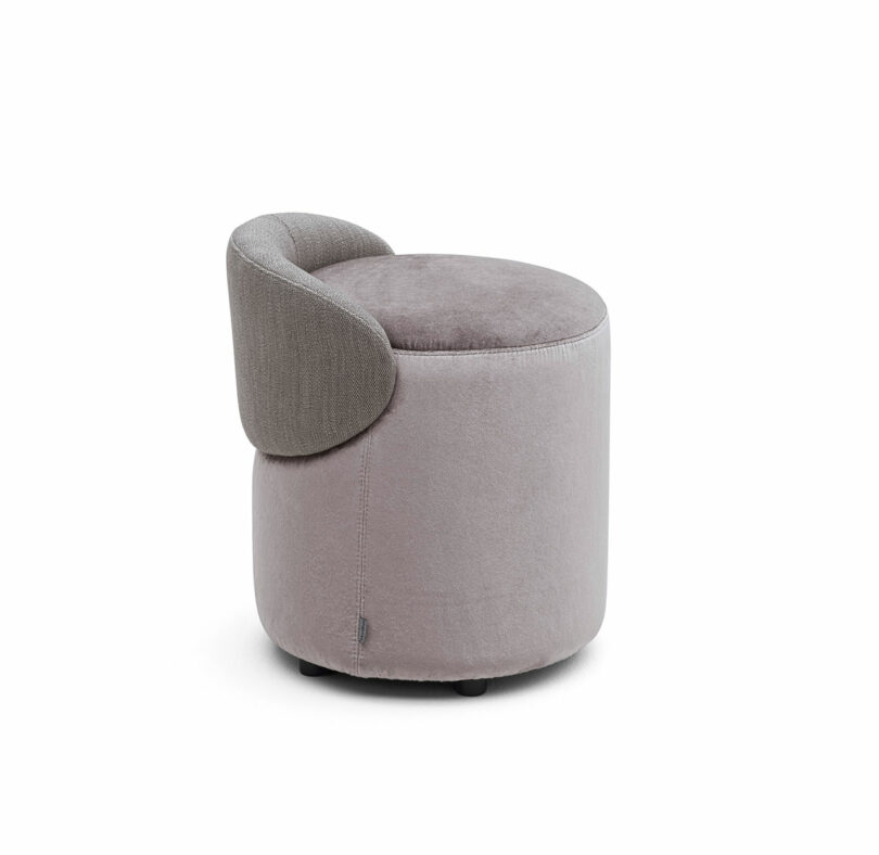 A round, light grey upholstered ottoman with a small, curved backrest in a slightly darker shade.