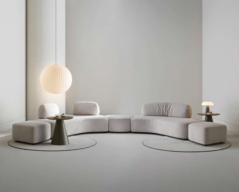 A modern living room with a light gray sectional sofa, two round side tables, a large hanging lamp, a small table lamp, and minimal decor on a light gray floor and walls.