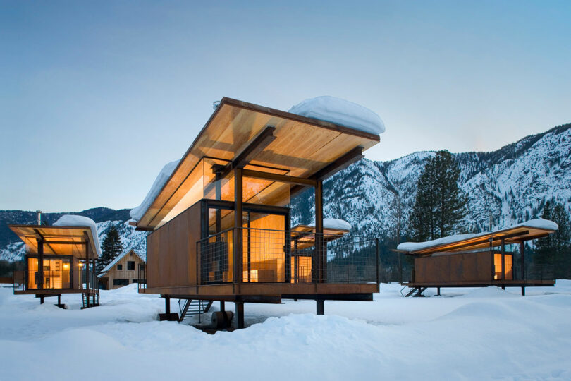Three modern wooden cabins on stilts sit in a snowy landscape with mountains in the background, illuminated from within at dusk.