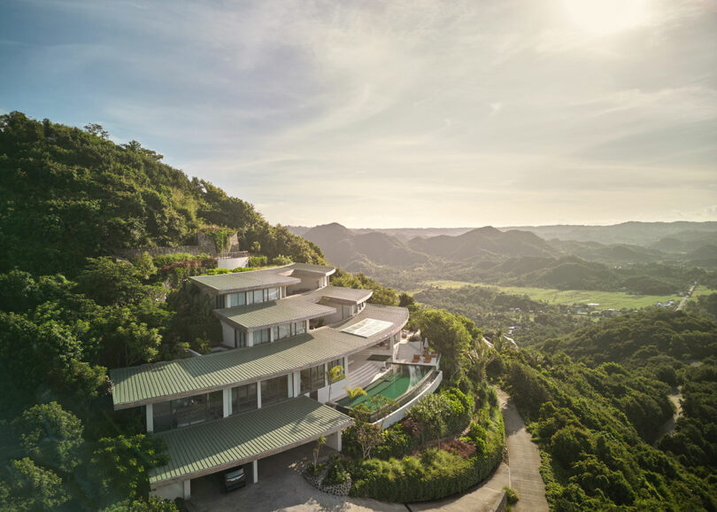 A modern multi-level house with a green roof sits on a hillside, overlooking a lush valley and distant mountains under a hazy sky.