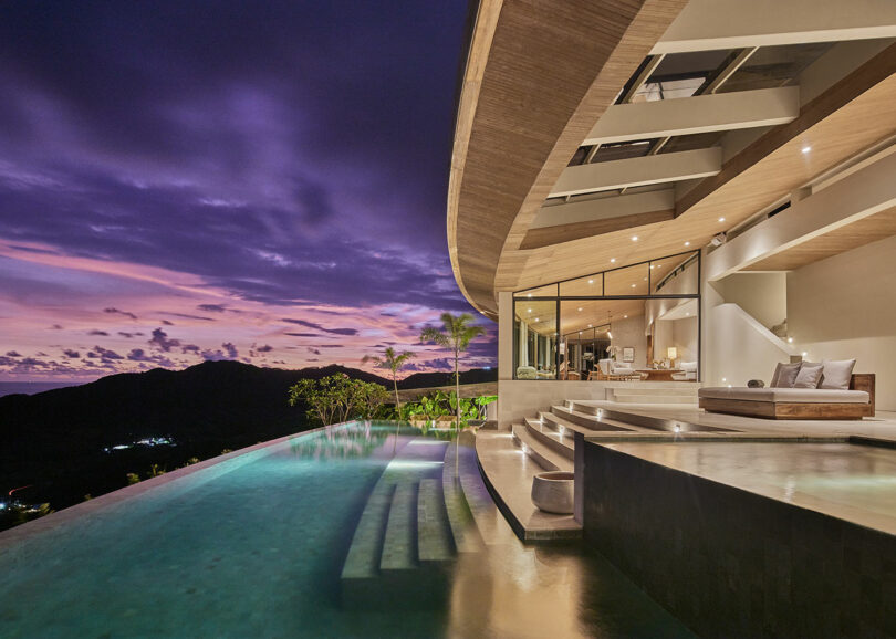 Modern luxury villa with open living area, infinity pool, and steps, overlooking mountains and a vibrant purple sunset sky.