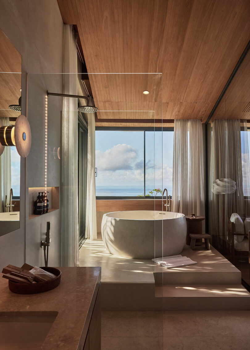 Modern bathroom with a large round stone bathtub, floor-to-ceiling windows, wooden ceiling, and an ocean view. Natural light fills the space.