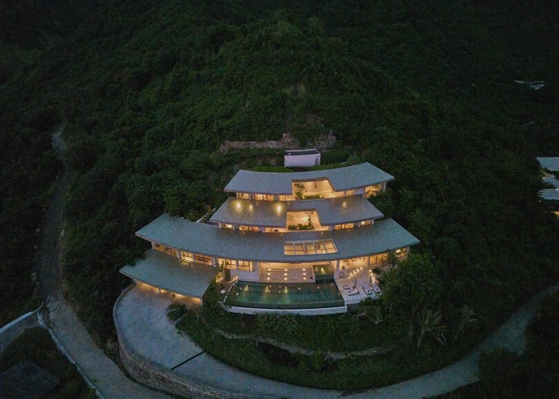 A large, multi-story modern house with illuminated windows is set on a lush, green hillside at dusk, with a pool and patio visible in front.