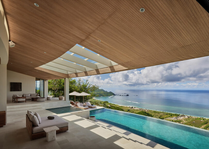 Infinity pool with lounge chairs under a wooden ceiling, overlooking a beach, ocean, and green hills under a partly cloudy sky.