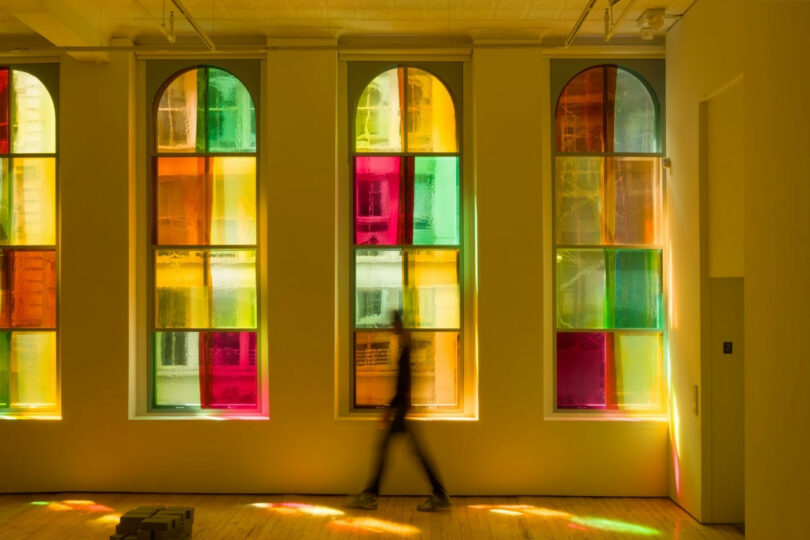 A person walks past three tall, arched windows with multicolored glass panels, casting colored light onto a wooden floor in this 2025 Unframed scene.