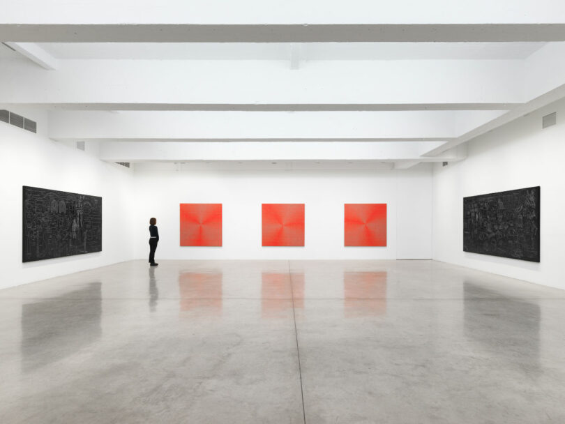 A person stands in a white-walled gallery viewing five abstract paintings, with three red geometric works on the back wall and two dark artworks on the side walls.