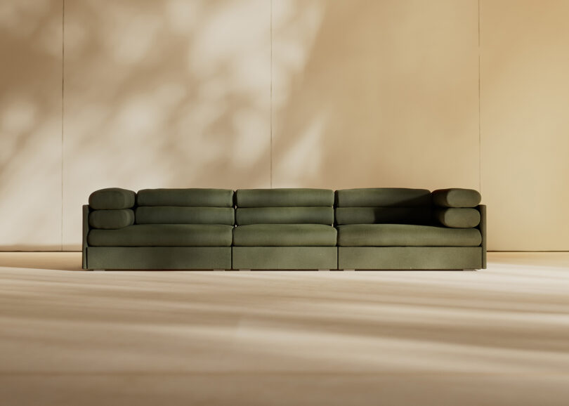 A modern, olive green modular sofa with cylindrical armrests is centered in a minimalist room with beige walls and soft, natural light casting shadows.