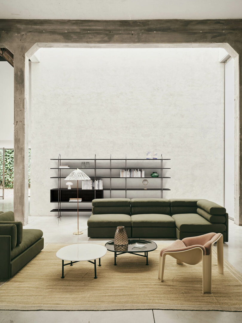 Modern living room with green sectional sofa, minimalist shelving, coffee tables, a floor lamp, and a beige chair, all set on a woven rug under a high ceiling with exposed concrete beams.