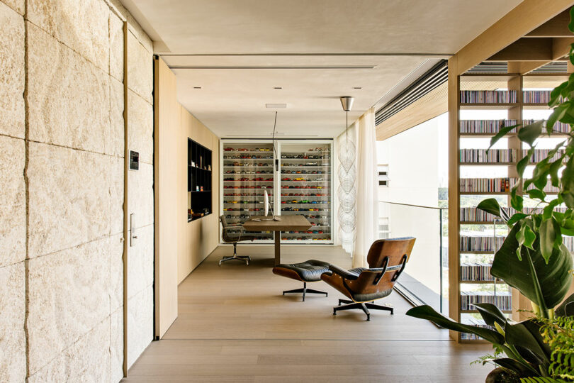 Modern office with a wall of model cars, an Eames lounge chair, built-in shelving, floor-to-ceiling windows, and indoor plants.