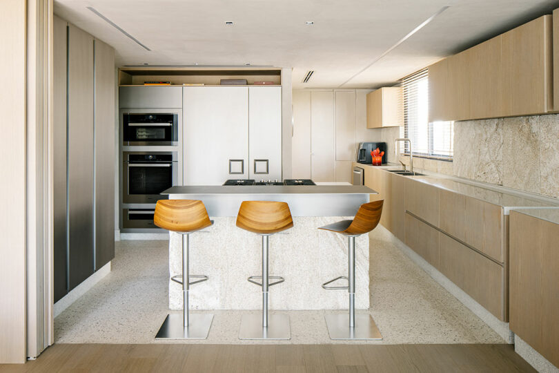 Modern kitchen with light wood cabinets, a central island with a marble countertop, three wooden bar stools, built-in ovens, and large windows with blinds.
