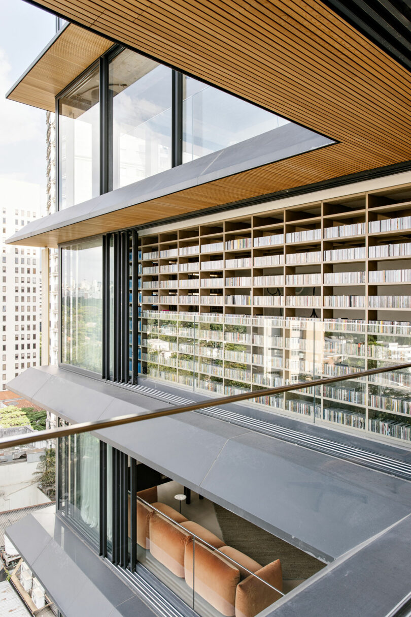 Modern building with large glass windows, horizontal metal louvers, and an interior lined with floor-to-ceiling bookshelves. A tan sofa is visible inside near the window.
