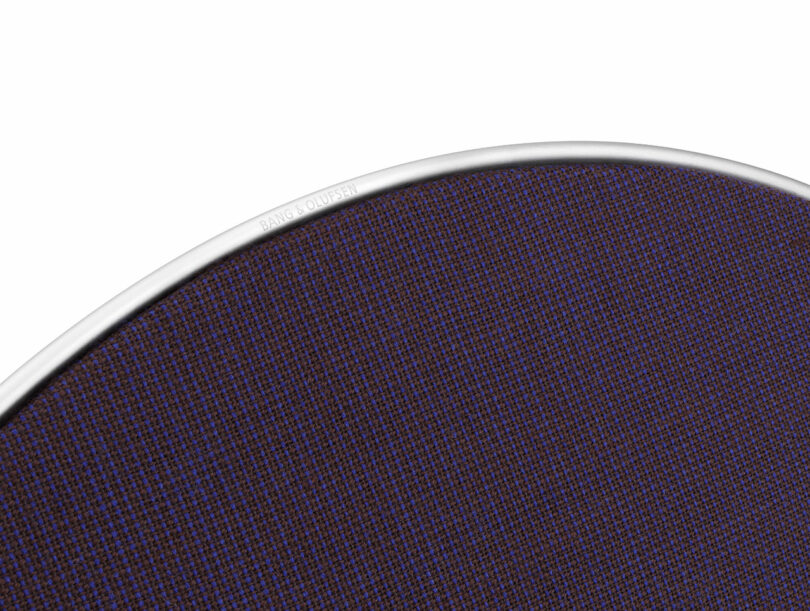Close-up of the edge of a round speaker with a dark purple mesh cover and a silver rim, branded with 