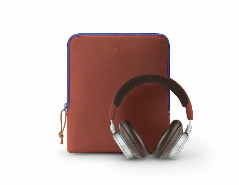 A brown tablet sleeve with a blue zipper stands upright behind a pair of brown and silver over-ear headphones.