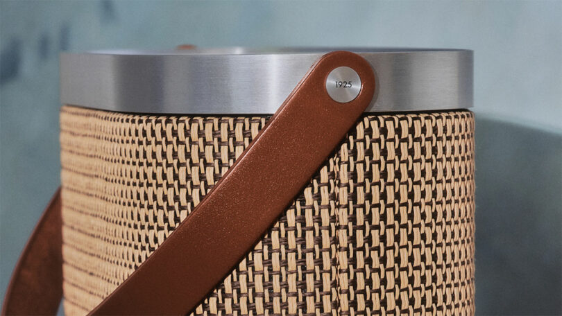 Close-up of a woven cylindrical object with a metal rim and a brown leather strap attached by a button engraved with 