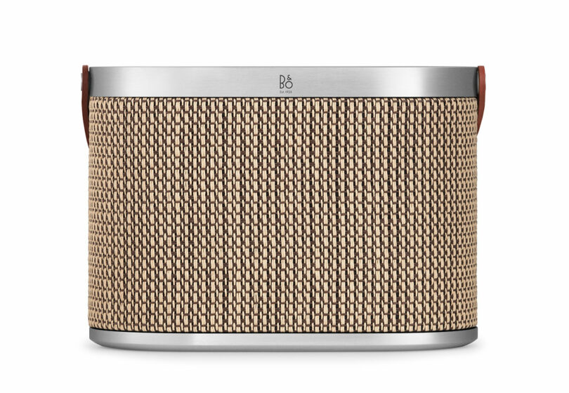 Rectangular wireless speaker with a woven tan and black front grille, silver metal top and bottom edges, and brown leather side handles.