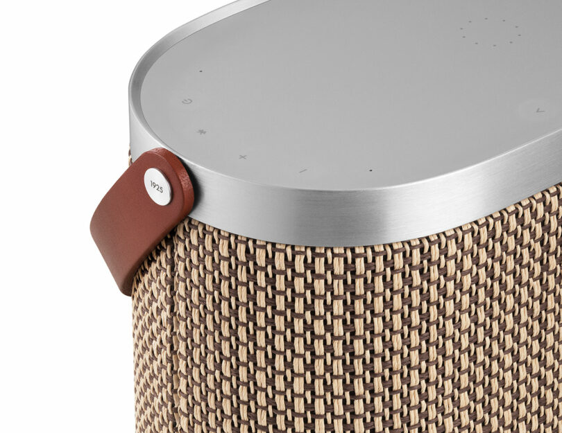Close-up of a modern wireless speaker with a woven fabric cover, metallic top panel, touch controls, and a brown leather strap with a metal tag.