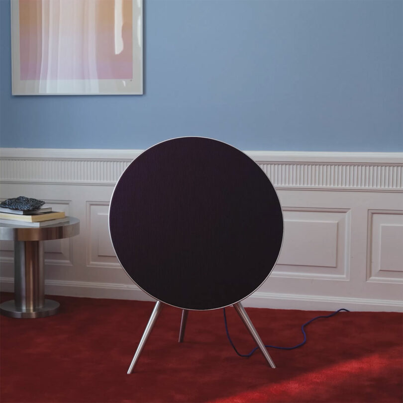 A round black speaker from the Centennial Collection with tripod legs stands on a red carpet in a room with blue walls, white paneling, and a framed abstract artwork.