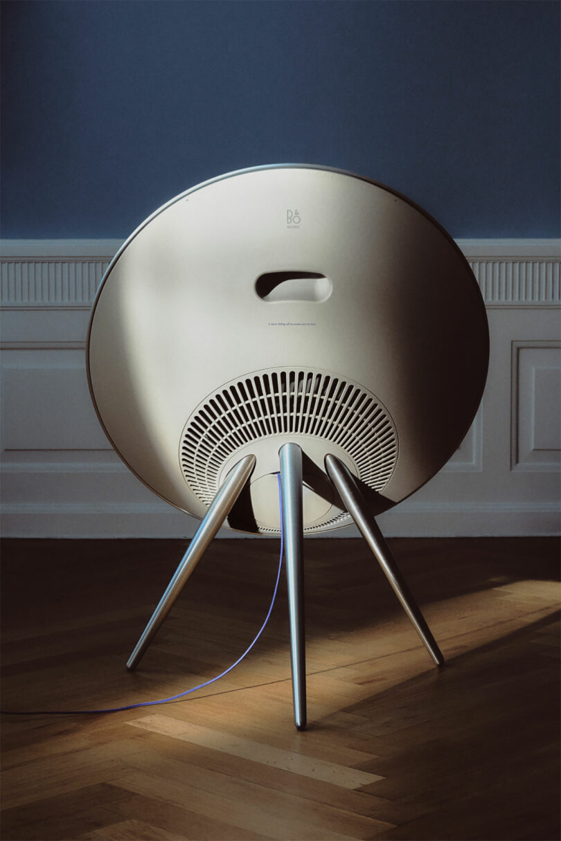 A circular speaker with tripod legs stands on a wooden floor against a white and blue wall, with cables extending from the back.