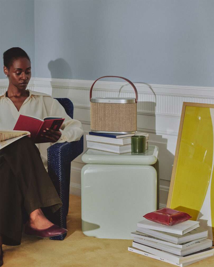 A woman sits in a blue chair reading a book beside a small table stacked with books, a bag, a candle, and a large yellow-framed artwork resting on the floor.