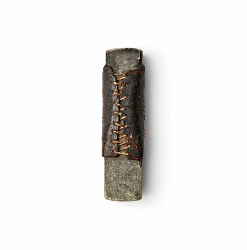 A Brent Buck stone tool with a rectangular blade and a leather cover stitched at the center, photographed on a white background.