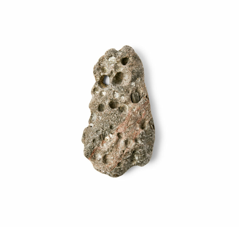 Irregularly shaped gray rock with multiple holes and a reddish-brown streak, reminiscent of a Brent Buck specimen, displayed on a white background.