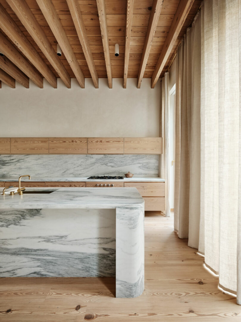 A modern Brent Buck kitchen features exposed wooden ceiling beams, light wood cabinetry, a marble island with matching backsplash, gold faucet, and floor-to-ceiling beige curtains.