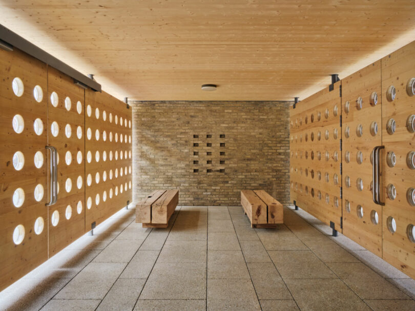 A Brent Buck-designed room with wooden walls and benches, featuring round cutouts and a brick wall at the end, with a tiled floor and ceiling lights.