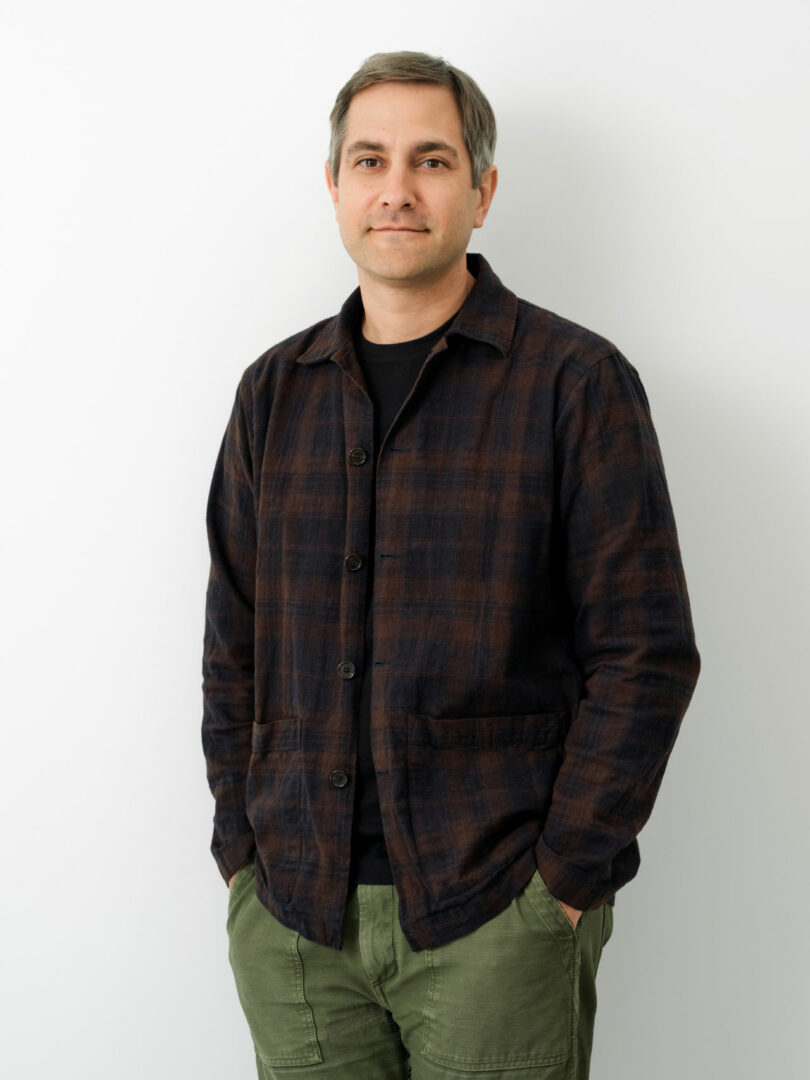 Brent Buck, a man with short hair, wears a dark plaid jacket over a black shirt and green pants as he stands against a plain white background with his hands in his pockets.