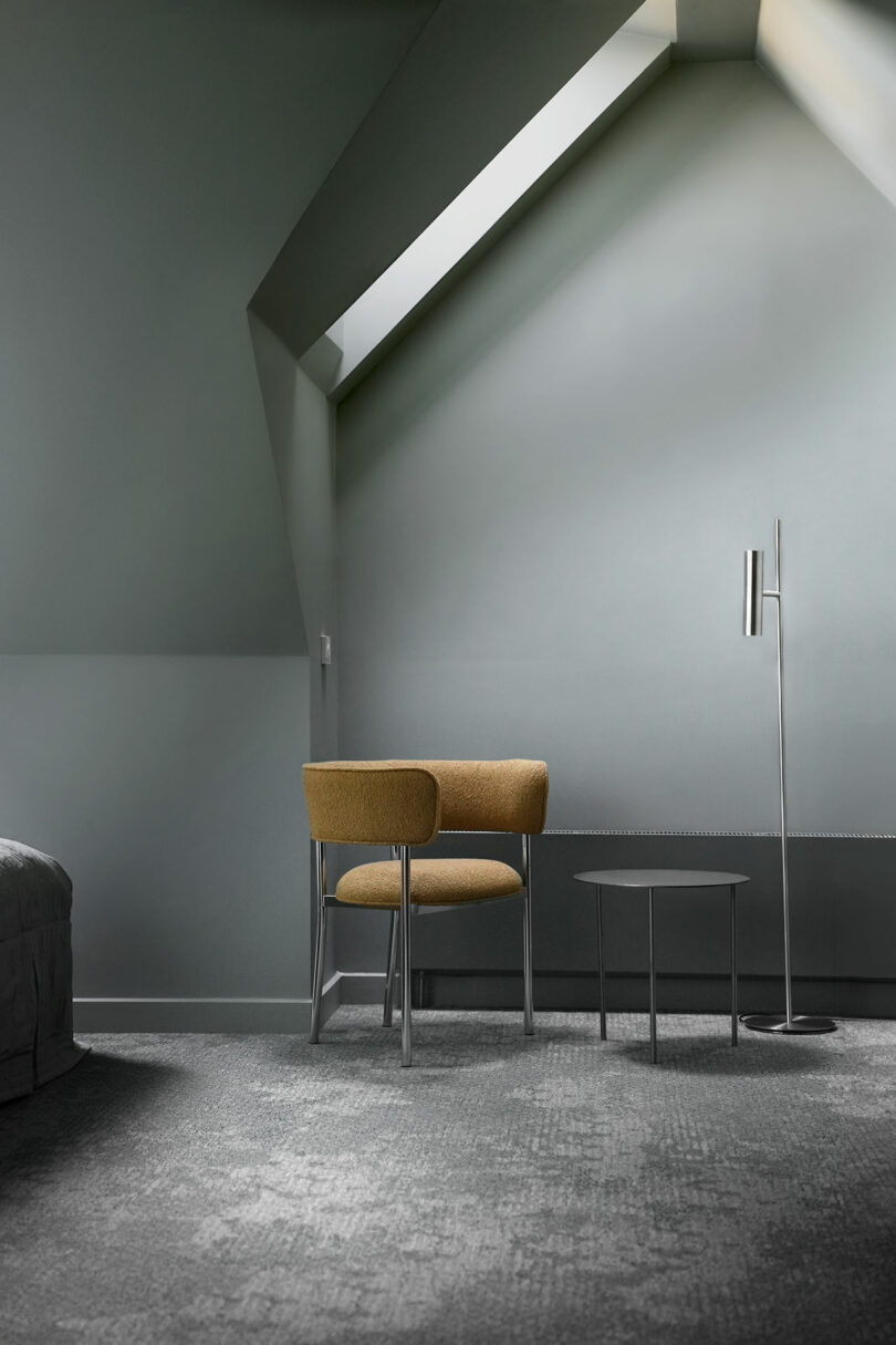 A modern, minimal room with a slanted ceiling and skylight, two tan upholstered chairs, a small round table, and a tall floor lamp on a grey carpeted floor.