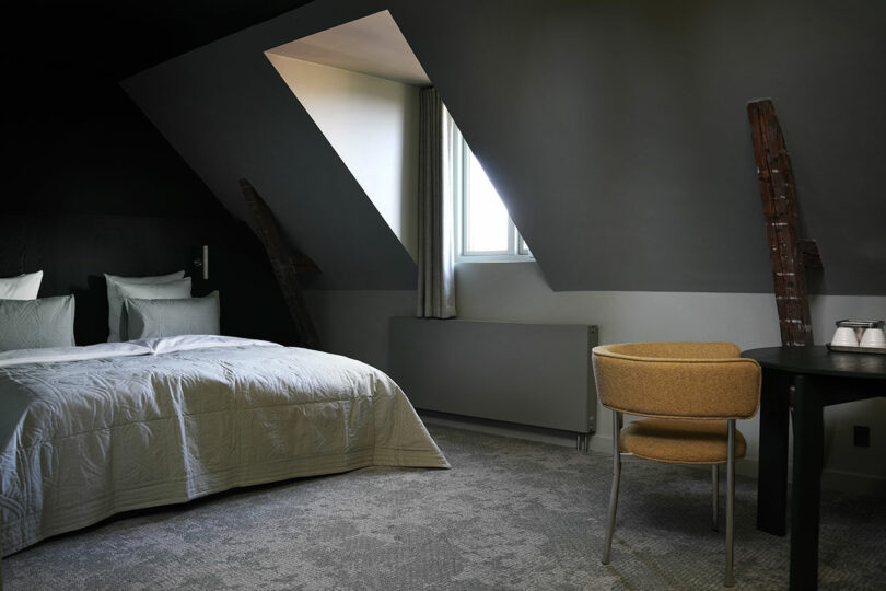 Modern hotel room with a large bed, angled ceiling with skylight window, exposed wooden beams, a round table, and a yellow chair.