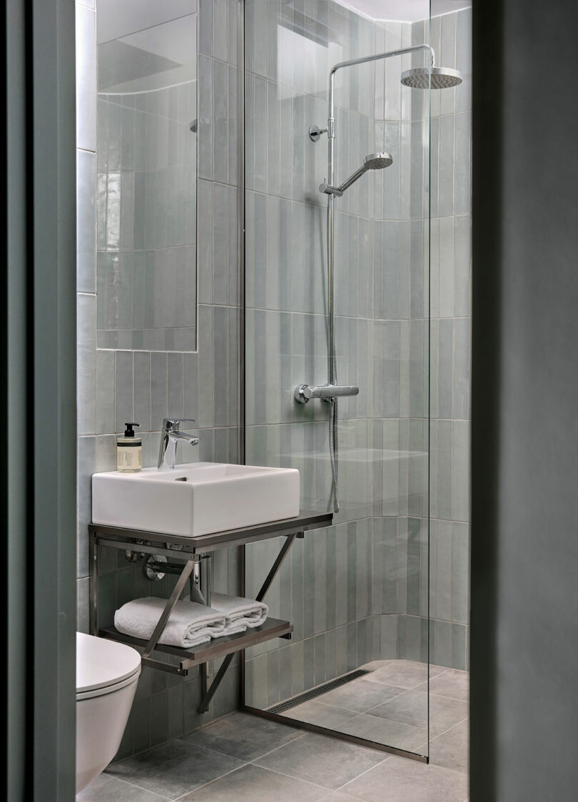Modern bathroom with a glass-enclosed shower, wall-mounted sink, folded towels on a shelf below, and a soap dispenser next to the faucet.
