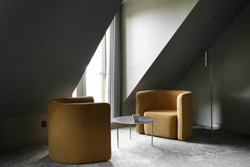 Two mustard yellow armchairs face a round black coffee table with a cup on it, set in a gray-carpeted room under a sloped ceiling with a tall window and floor lamp.