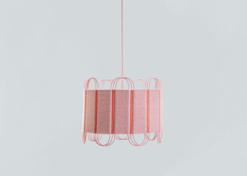 A pink pendant light with a cylindrical shade wrapped in pink cord and a decorative wire frame, designed by Laura Casañas Maya, hanging against a plain light gray background.