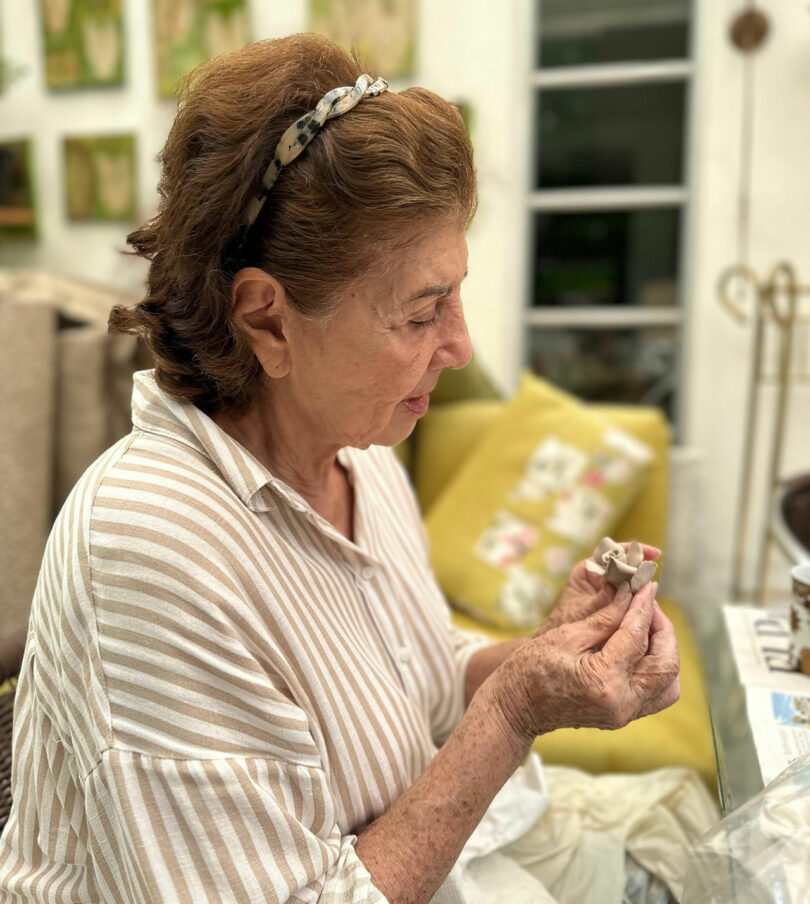 An older woman in a striped shirt sits indoors, closely examining a small clay object in her hands, reminiscent of the intricate work by Laura Casañas Maya.
