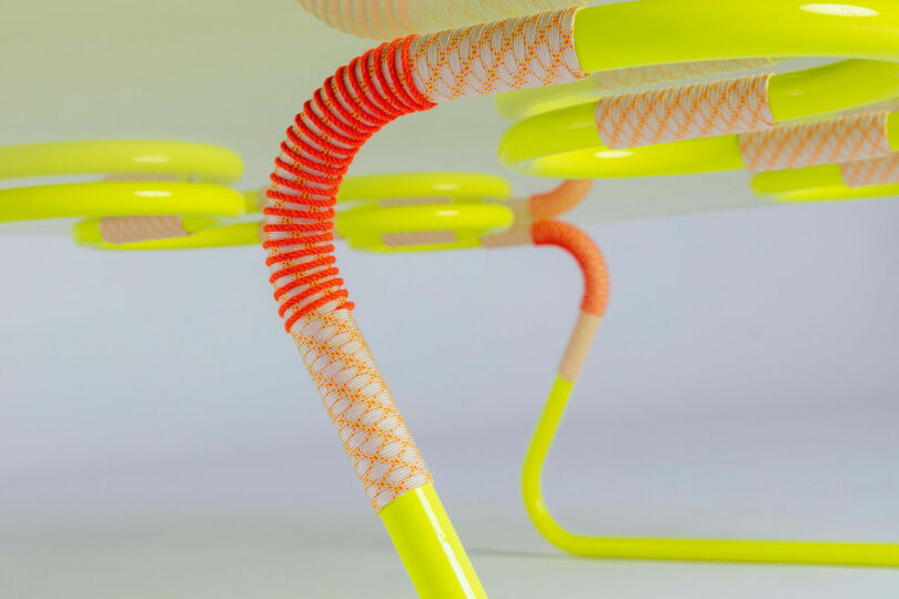 Close-up view of a table by Laura Casañas Maya, featuring yellow metal legs wrapped in orange and white textured cord, set against a plain background.