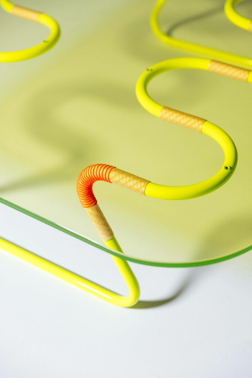 Close-up of a glass table with yellow, curvy metal legs partially wrapped in orange and beige cord, designed by Laura Casañas Maya.