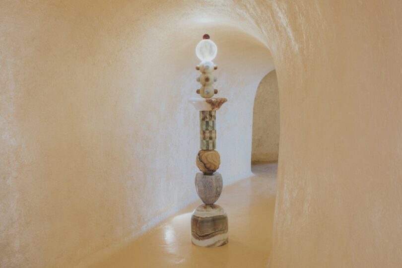 A tall, abstract stone sculpture stands in a curved, beige-toned hallway with smooth, textured walls and an arched doorway in the background