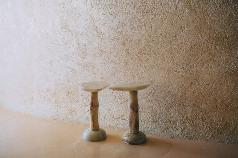 Two small, round stone stools with uneven tops stand side by side against a textured, beige plaster wall